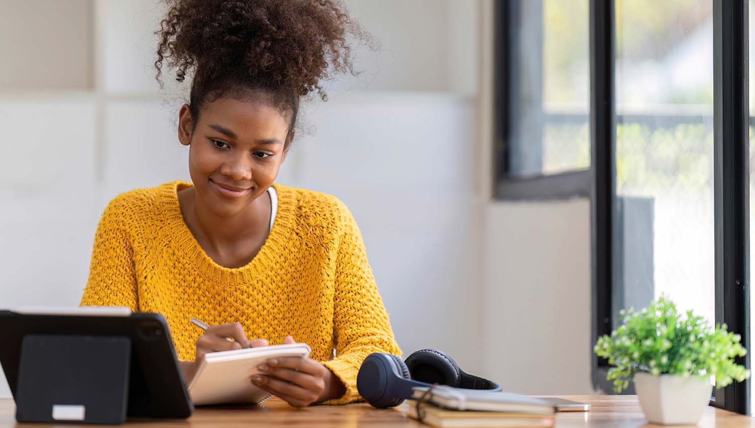 Student wearing a yellow sweater writing notes while using a tablet at a desk