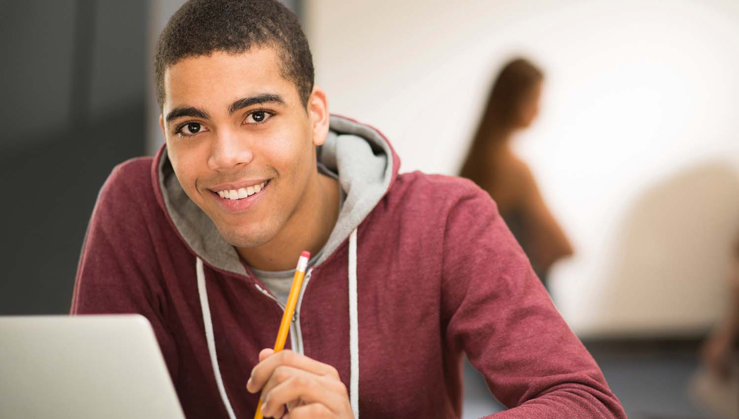 Smiling student in a red hoodie working on a laptop while holding a pencil